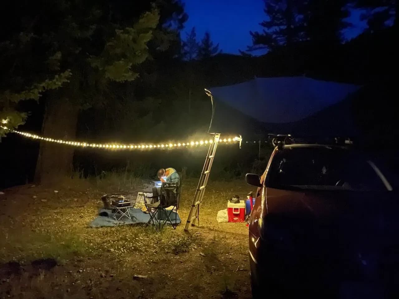 Campo di notte con lucine, sedie da campeggio e l'auto parcheggiata sotto gli alberi