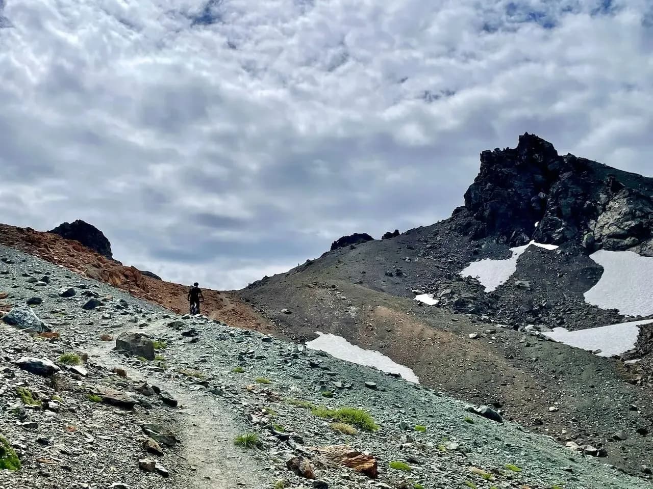 Singletrack che attraversa un ghiaione con chiazze di neve sotto una cima rocciosa