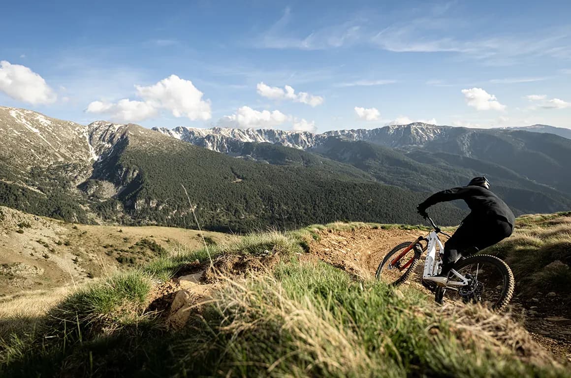 Rider en e-bike por un sendero de tierra entre prados alpinos de Grandvalira con picos nevados al fondo
