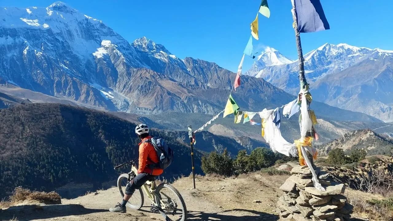 Rider parado junto a banderas de oración en un sendero de Mustang, frente a cumbres nevadas del Himalaya