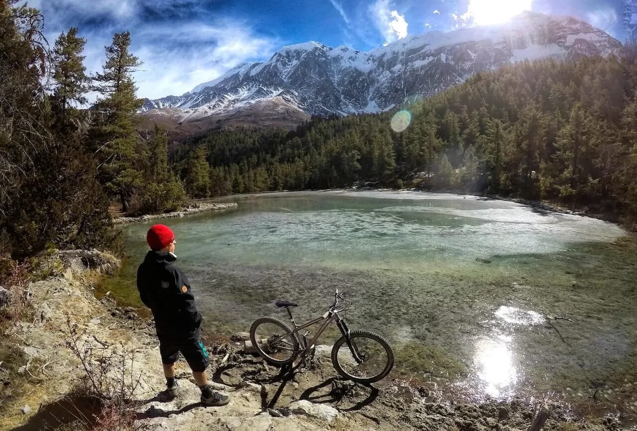 Rider con gorro rojo junto a su bici a orillas de un lago glaciar de Mustang, con montañas nevadas al fondo