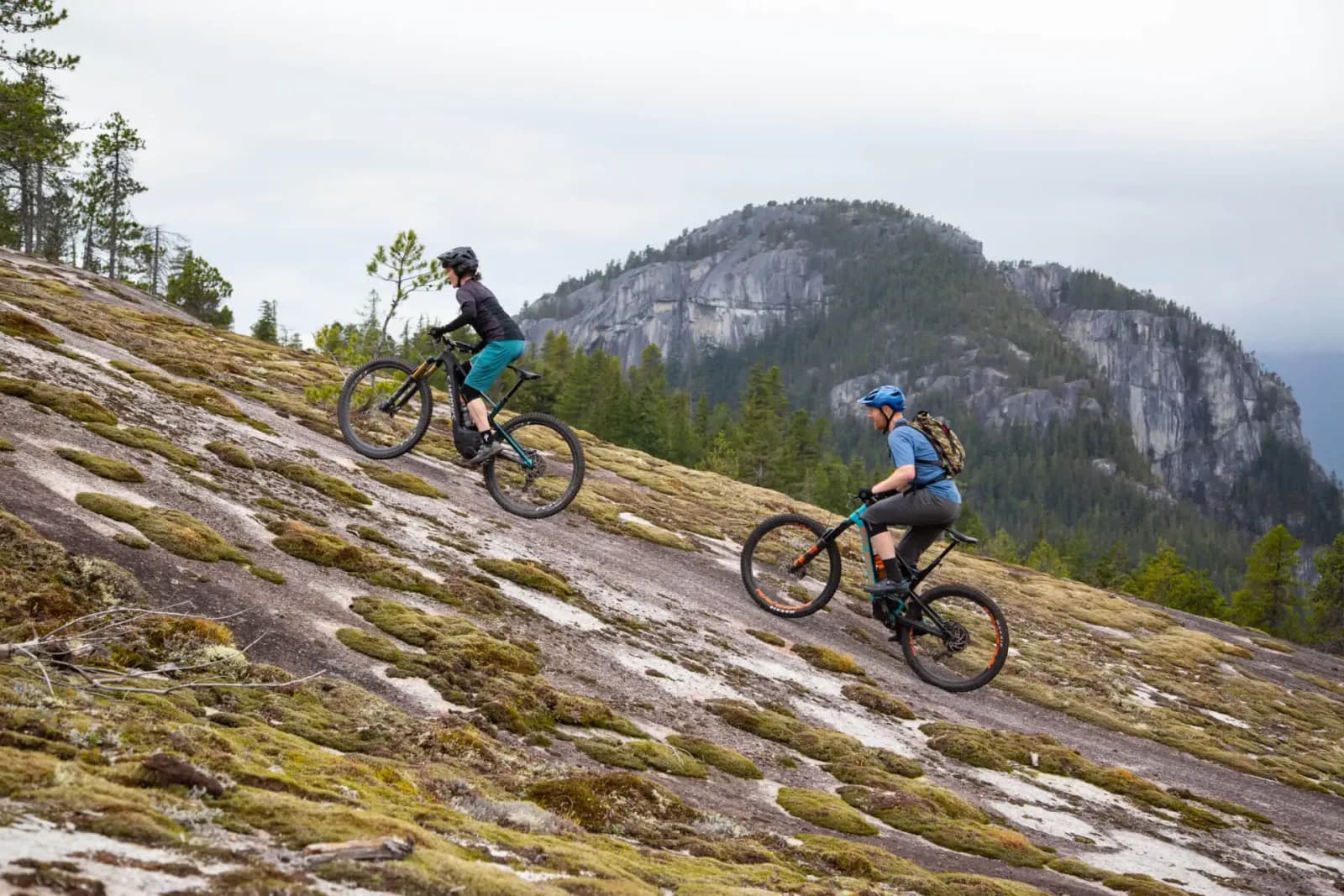 Zwei Rider fahren mit E-Bikes eine moosbewachsene Granitplatte hinauf, dahinter Felswände in Kanada