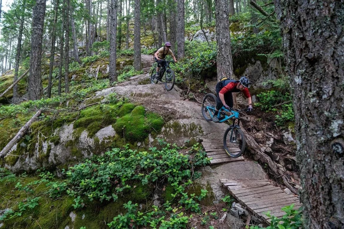 Zwei Rider fahren in einem Wald in Squamish über eine Granitplatte und einen Holzsteg hinunter