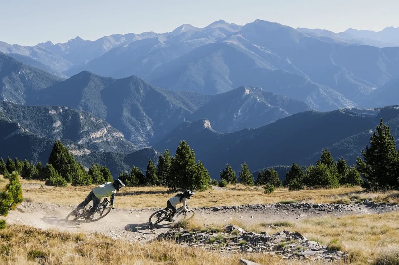 Zwei Rider in einer Kurve auf einem staubigen Trail in Vallnord Pal-Arinsal, im Hintergrund die Pyrenäen