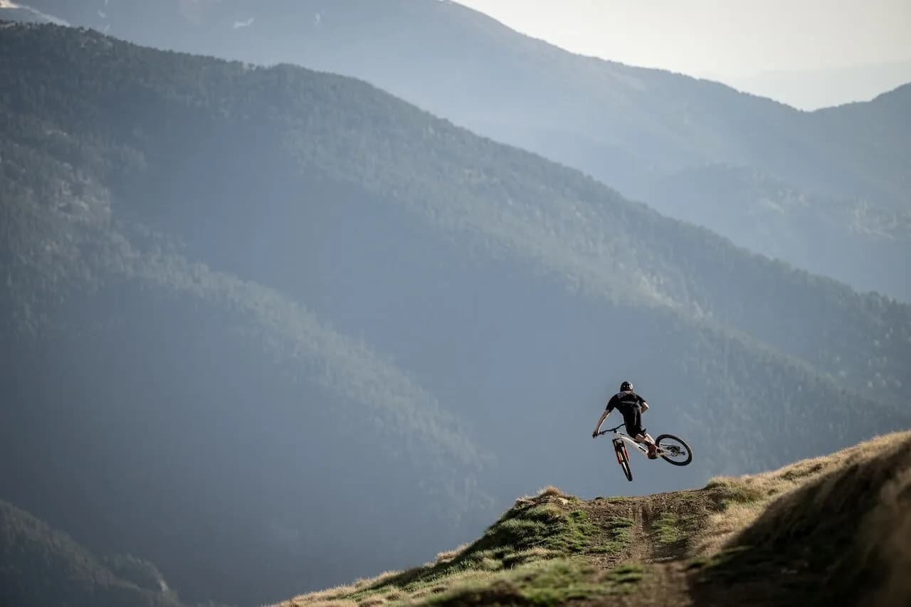 Rider springt über einen grasbewachsenen Kamm, dahinter die bewaldeten Berge Andorras
