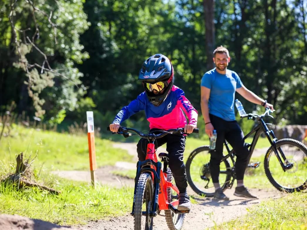 Kind mit Fullface-Helm, Goggle und langärmligem Trikot fährt in einem Bike Park neben einem Erwachsenen mit seinem Bike